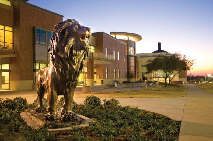 Lion statue in front of a bricked building. It is in the evening and the focus is on the lion.