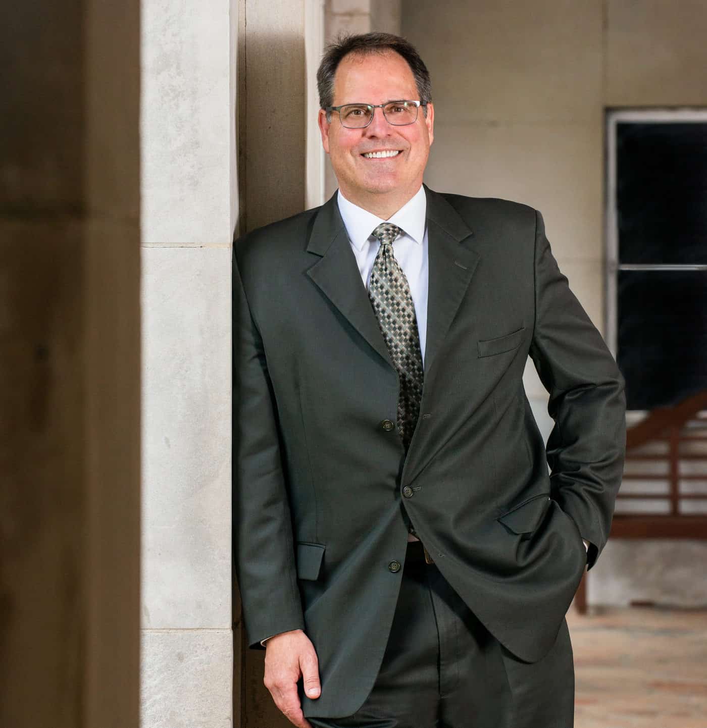A smiling man in a dark suit leans casually against a stone pillar, posing in a formal yet relaxed manner.
