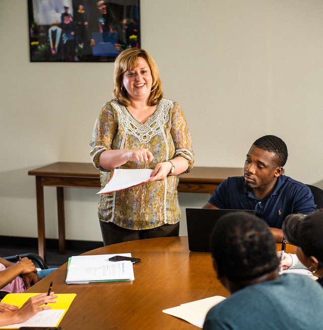 Woman holding a file while speaking to group around a conference table.