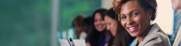 Mid adult African American businesswoman is smiling and looking at the camera. She is using her smart phone to take notes while listening to a speaker during a business conference or job training seminar. She's sitting in a lecture hall with professional colleagues.