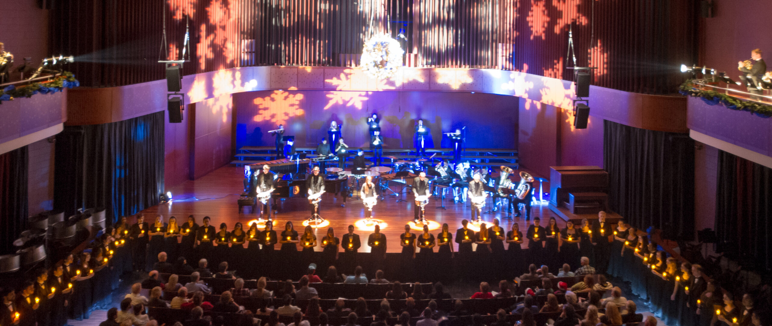 A holiday-themed performance on stage with musicians and singers holding candles, with snowflake lights projected in the background.