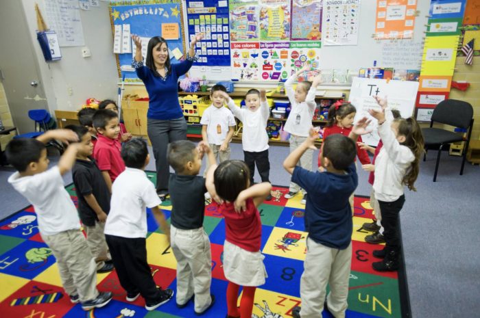A teacher stands in front of a group of young students in a colorful classroom. The children are excitedly participating in an interactive lesson, raising their arms while standing on a vibrant alphabet rug.