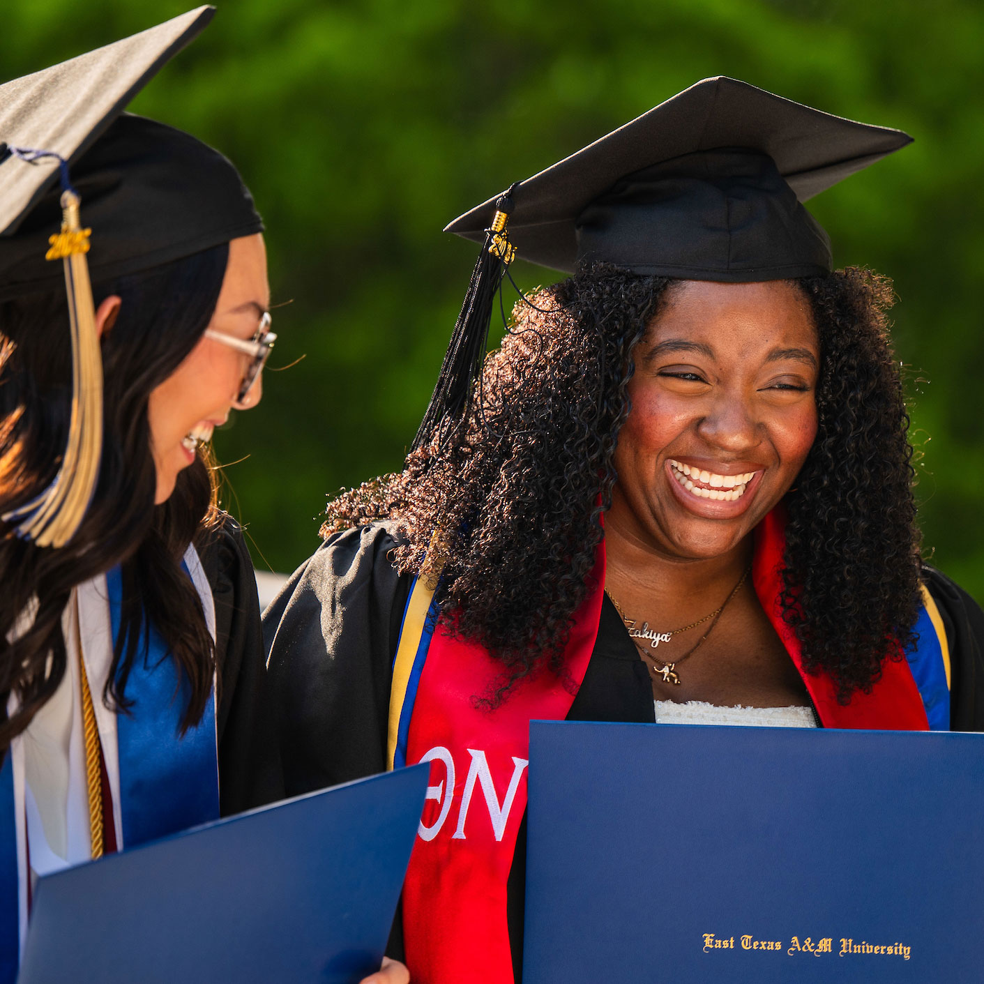 Two students in graduation cap and gown smile holding their diploma.