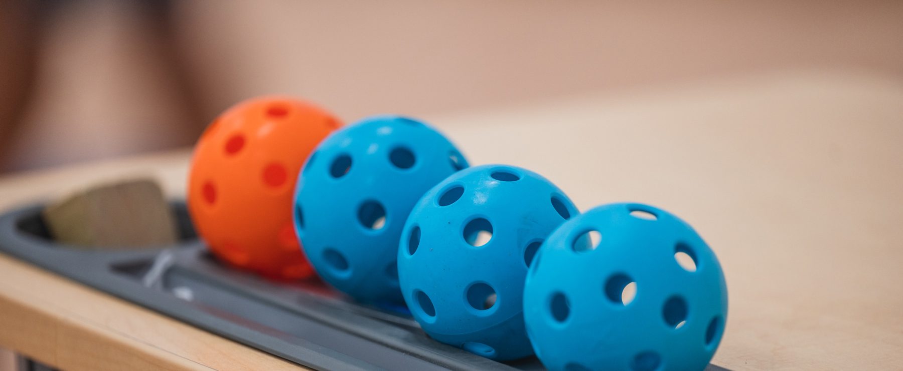 Four perforated plastic balls, two blue and two orange, are lined up in an orderly fashion on a beige tabletop. The balls rest in a grey tray, and their design suggests they are for a sport or game such as pickleball. The background is blurred.