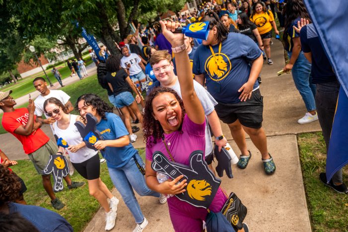 A group of excited students celebrates during a campus event, one raising a foam finger and cheering while wearing a purple shirt, with others smiling and holding Lion-themed props.