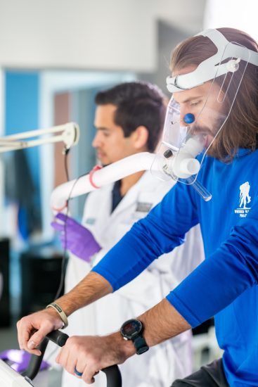 A man in a blue shirt cycles on an exercise bike wearing a breathing mask, monitored by a researcher in a lab coat. The setting is a scientific or medical research facility.