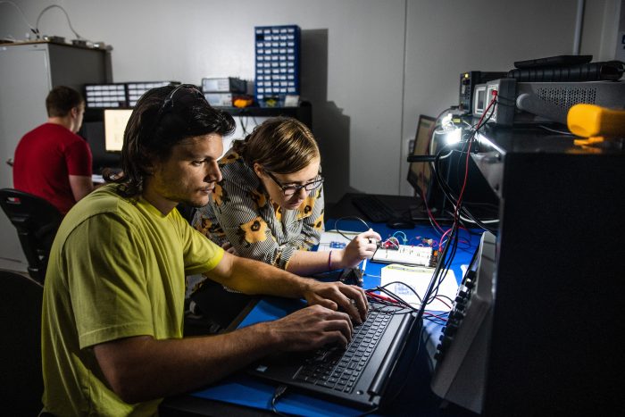 Two individuals are working together at a desk equipped with electronic components and a laptop. The man uses the laptop, while the woman interacts with electronic devices and wires while looking at the computer screen. Another person is in the background focusing on their computer. The room is dimly lit.