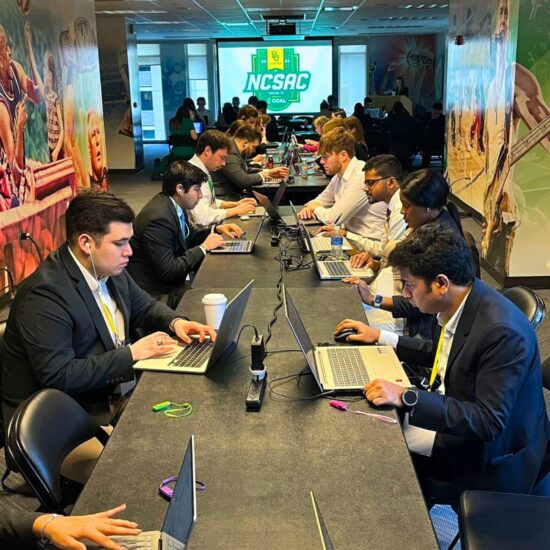 A group of individual sitting at a long desk with their laptops working.