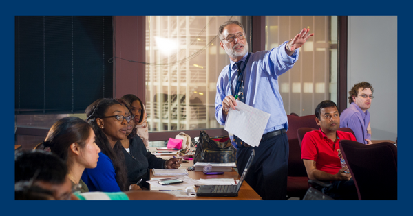 A professor gestures while holding a paper, addressing attentive students seated around a classroom table. The scene reflects a focused and engaged learning environment.