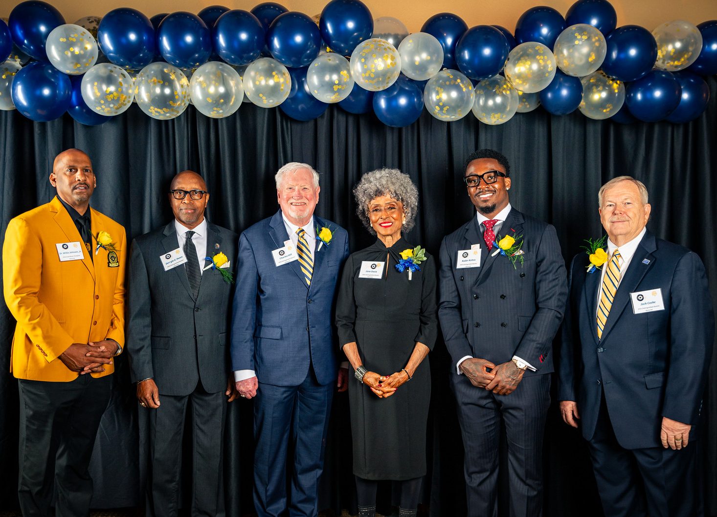 Six awardees stand side-by-side for a photo in front of a black curtain with blue and white balloons overhead.