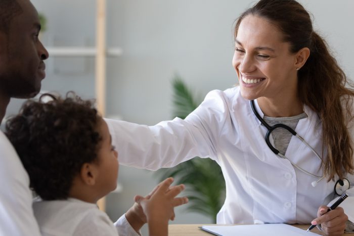 Friendly female doctor stroke head of cute african child boy patient sit on dads lap, black father with child visit pediatrician show good attitude to trusting child, children health care concept