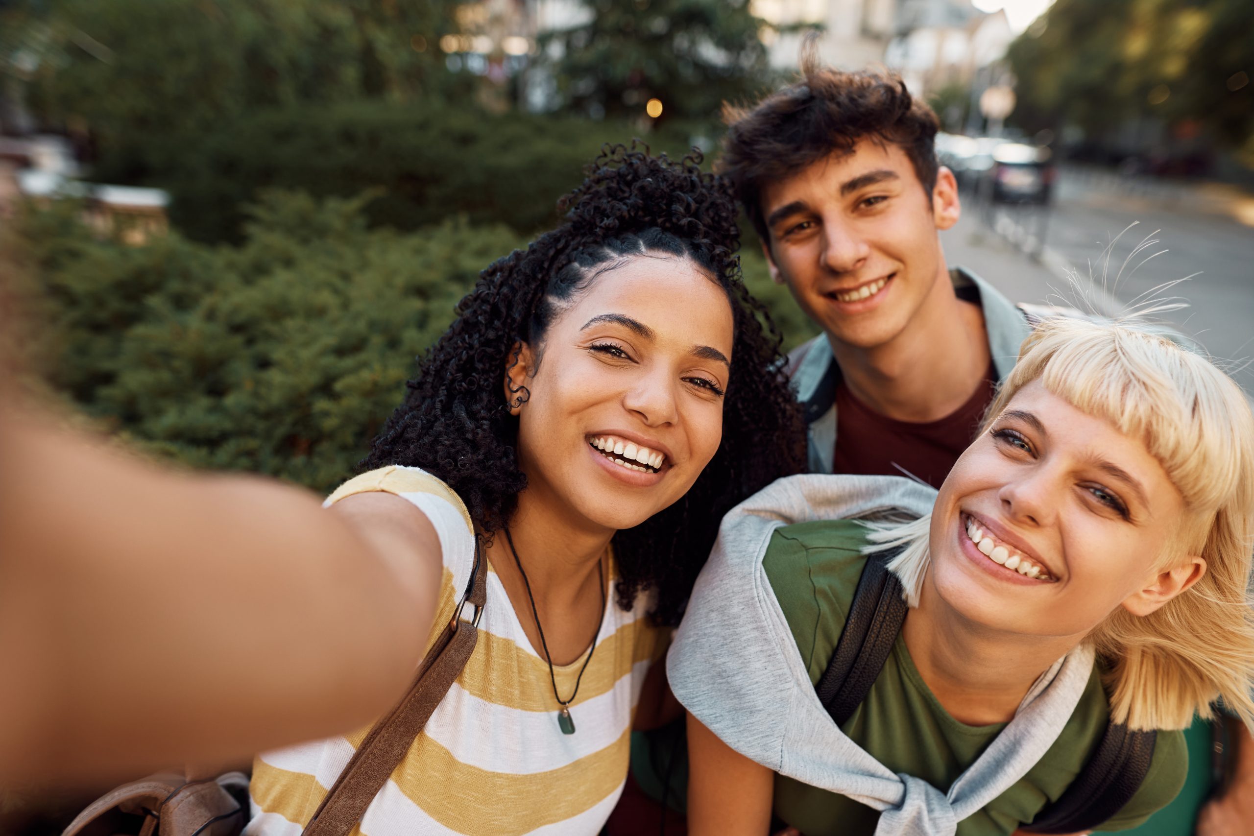Three friends smile and pose for a cheerful selfie outdoors, capturing a moment of friendship and joy in a green, urban setting.