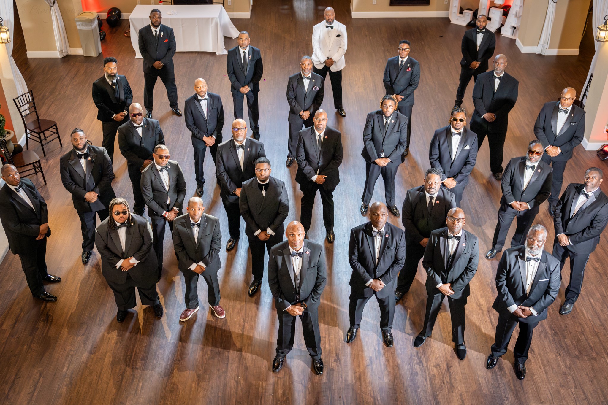 A formal group photo showing a gathering of men in black tuxedos, standing in a semi-circle on a wooden floor, with one individual in a white tuxedo at the top center.