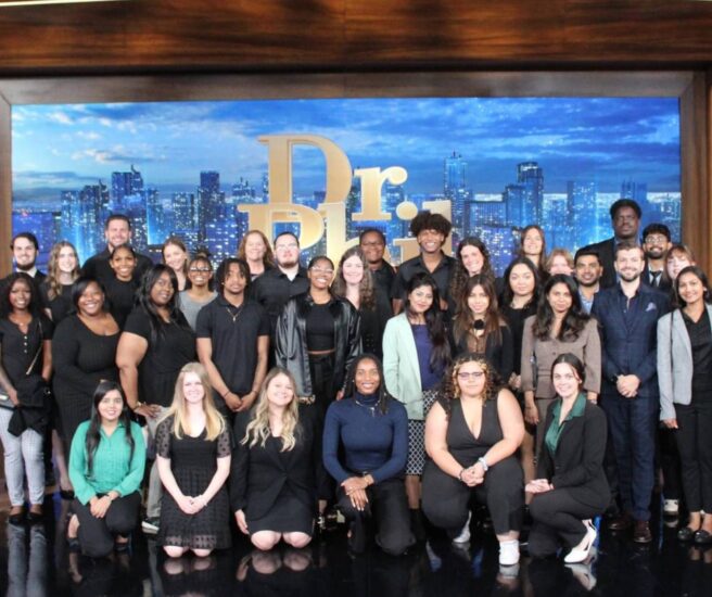 A group of people posing together on a stage in front of a backdrop with the "Dr. Phil" logo and a cityscape.