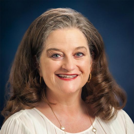 Portrait of a smiling woman with long curly hair, wearing a white blouse and gold earrings. Professional headshot with a dark blue background.
