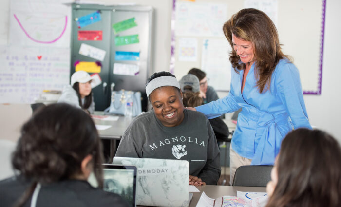 A teacher in a blue blouse smiles and places a hand on a student's shoulder. The student, wearing a gray "Magnolia" sweatshirt, smiles at her laptop. Other students work in the background.