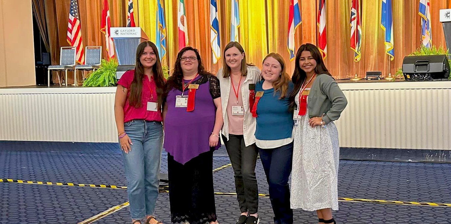 Four smiling female college students pose with a female faculty member in the middle. A stage behind them is adorned with flags representing countries around the world.