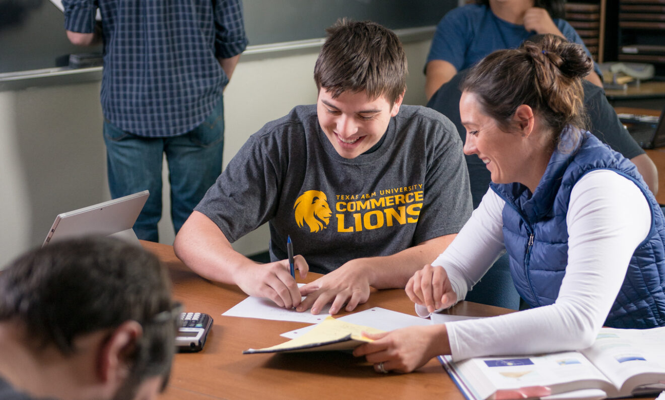 Two students work together at a table in a classroom. A student in the background is at the blackboard.