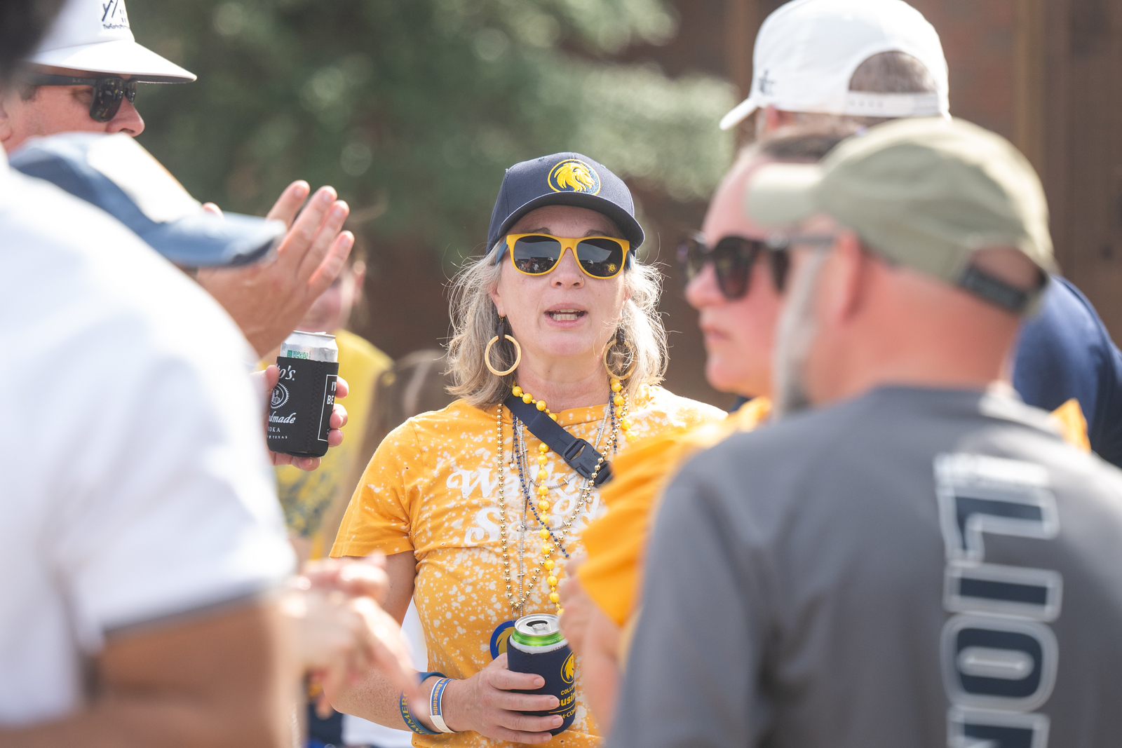 A woman wearing a yellow "Walking Strong" shirt, yellow sunglasses, and a cap with a lion logo is socializing outdoors with a group of people. She is holding a can with a beverage koozie, and others around her are casually conversing in the sunlight.