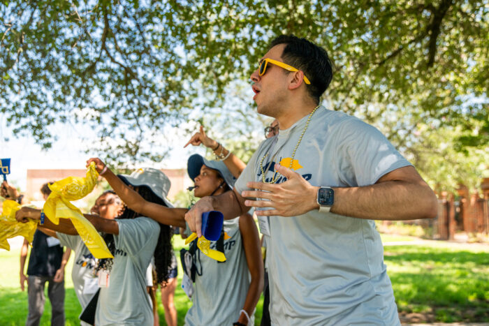 A group of enthusiastic students at an outdoor event cheer on their peers, led by a participant wearing sunglasses and holding a bandana, creating an energetic and spirited atmosphere.