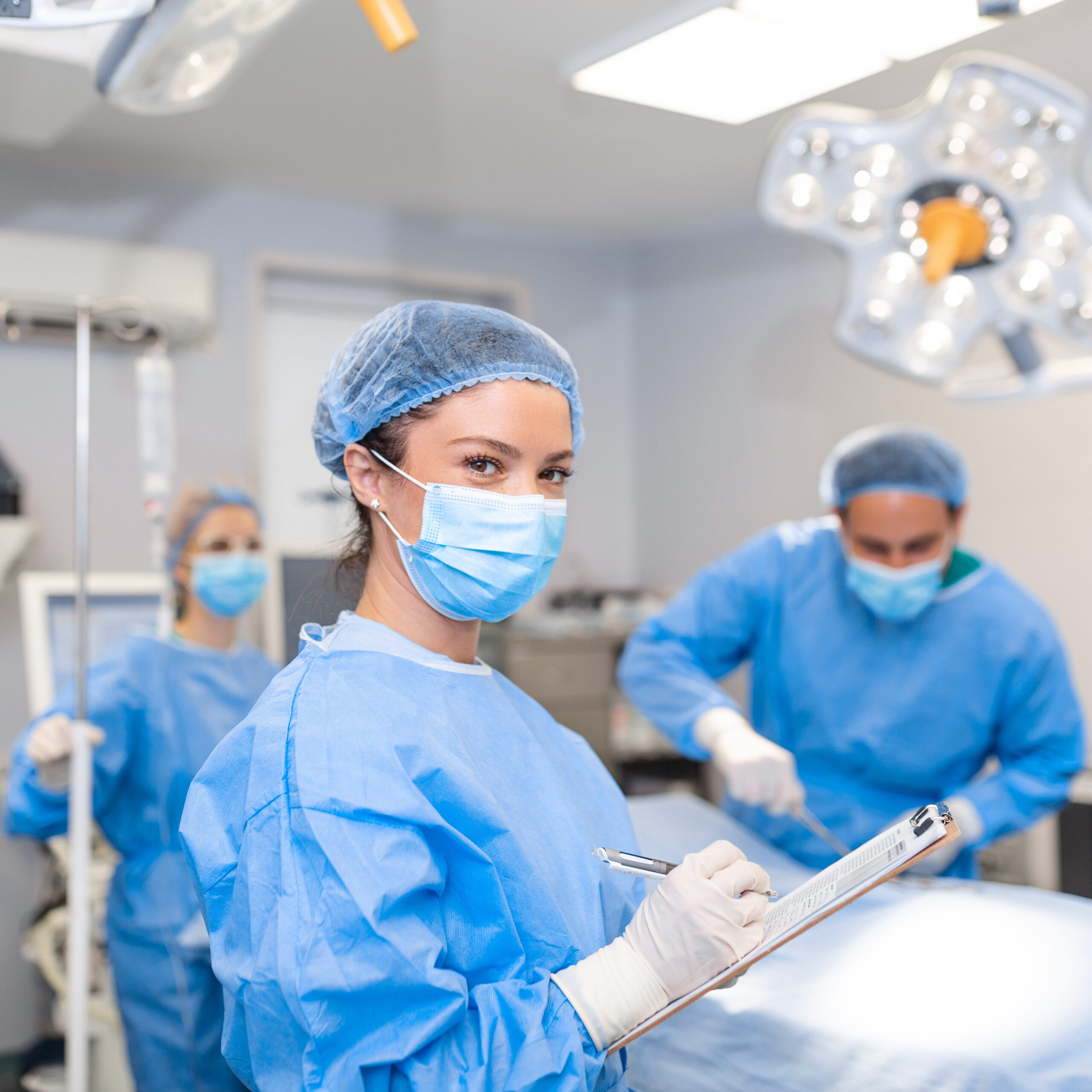 Woman wearing blue surgical gown and mask, holding clipboard in blue room with medical staff.