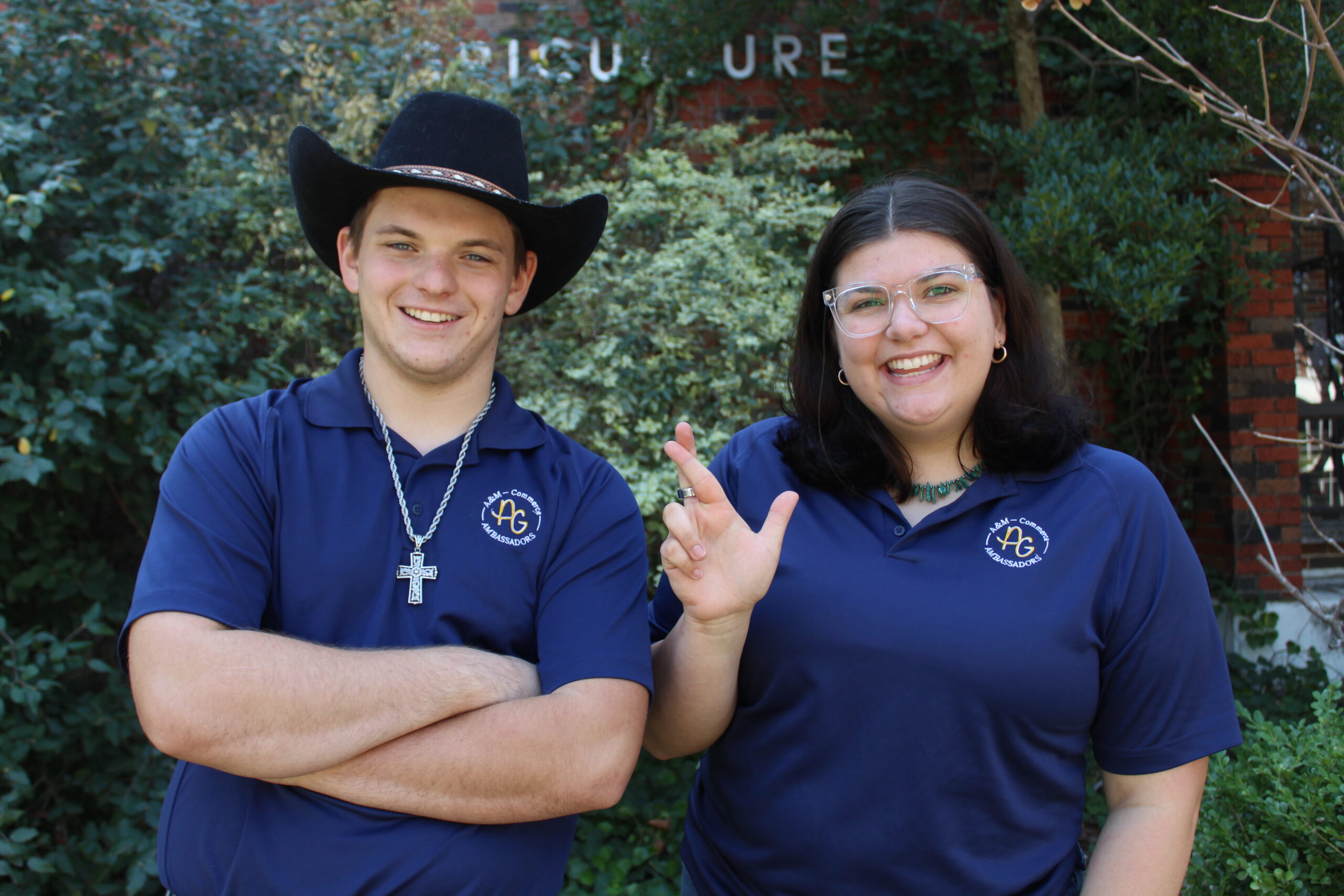 Two people wearing matching blue polo shirts stand outside in front of greenery. The man on the left wears a black cowboy hat and cross necklace, while the woman on the right is smiling and making the lion hand gesture.