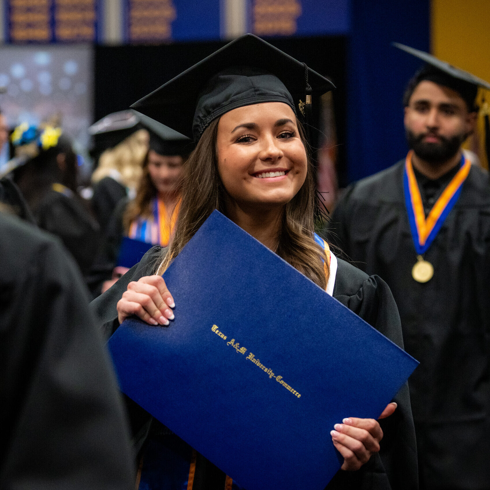 Graduate holds diploma in both hands and smiles at the camera, wearing graduation cap and gown.
