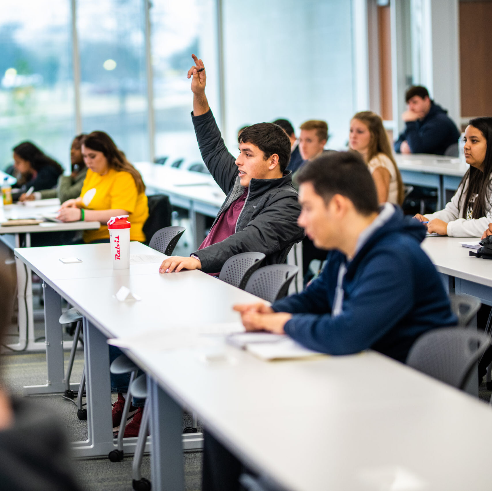 A classroom full of students, one of them with his hand raised to ask a question.