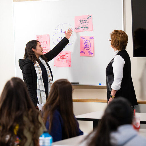 Female student presenting in front of class at a white board with female teach watching.