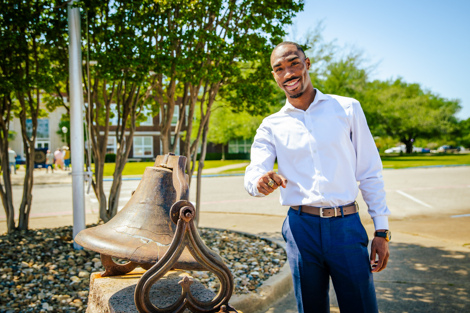 A young black male wearing a white button shirt with blue dress pants is standing by a monument of a bell. He is gesturing with his hands.