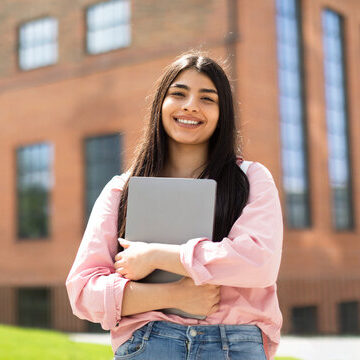 Portrait of happy lady student posing with laptop in hands outdoors, looking and smiling at camera