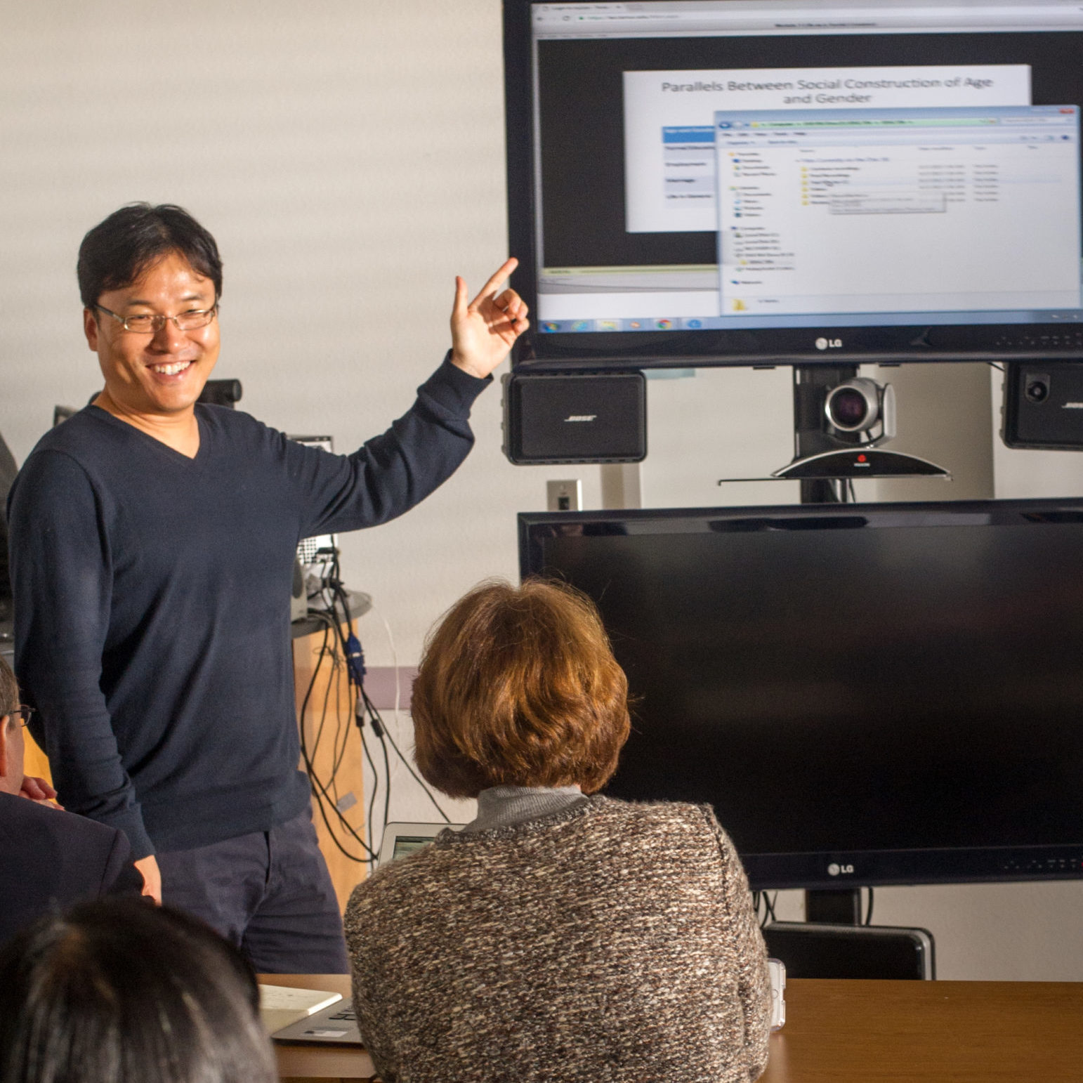 Male teacher point at TV screen and conferencing camera in front of class.