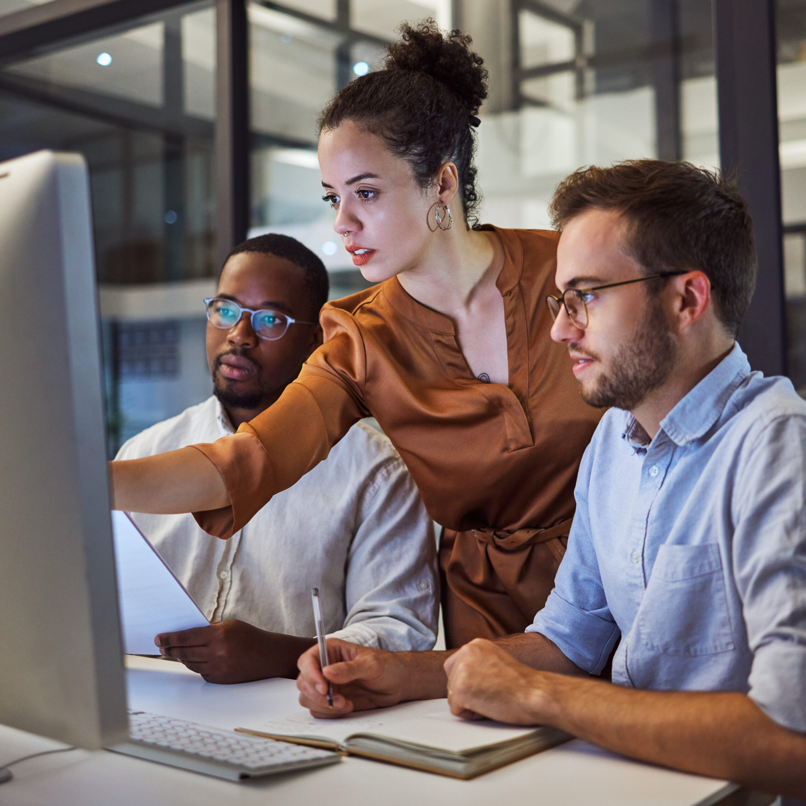 Three individual in front of a computer. One of them is pointing to the screen while the other two appear to be listening.