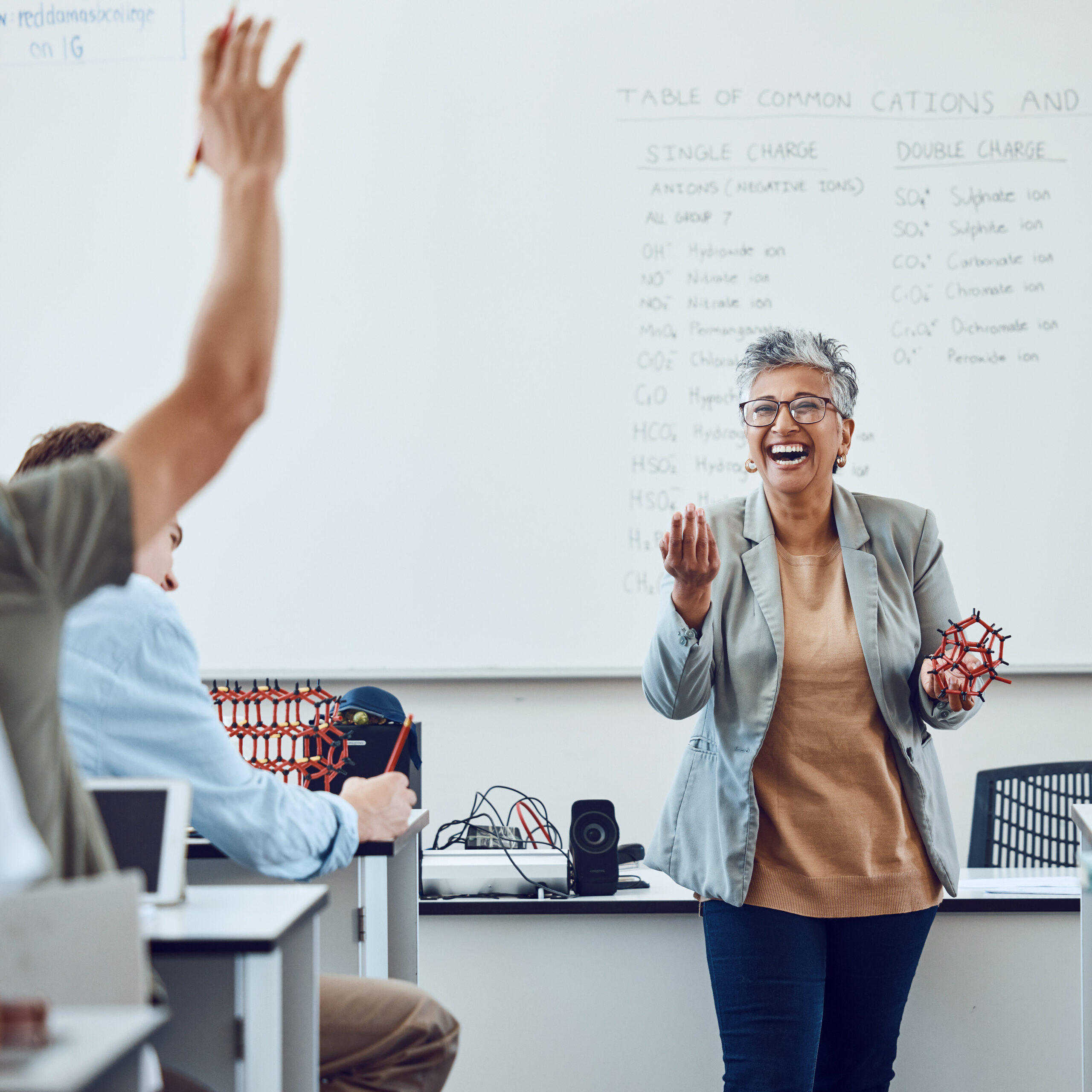 Teacher lecturing in front of a full classroom of students with hands raised.