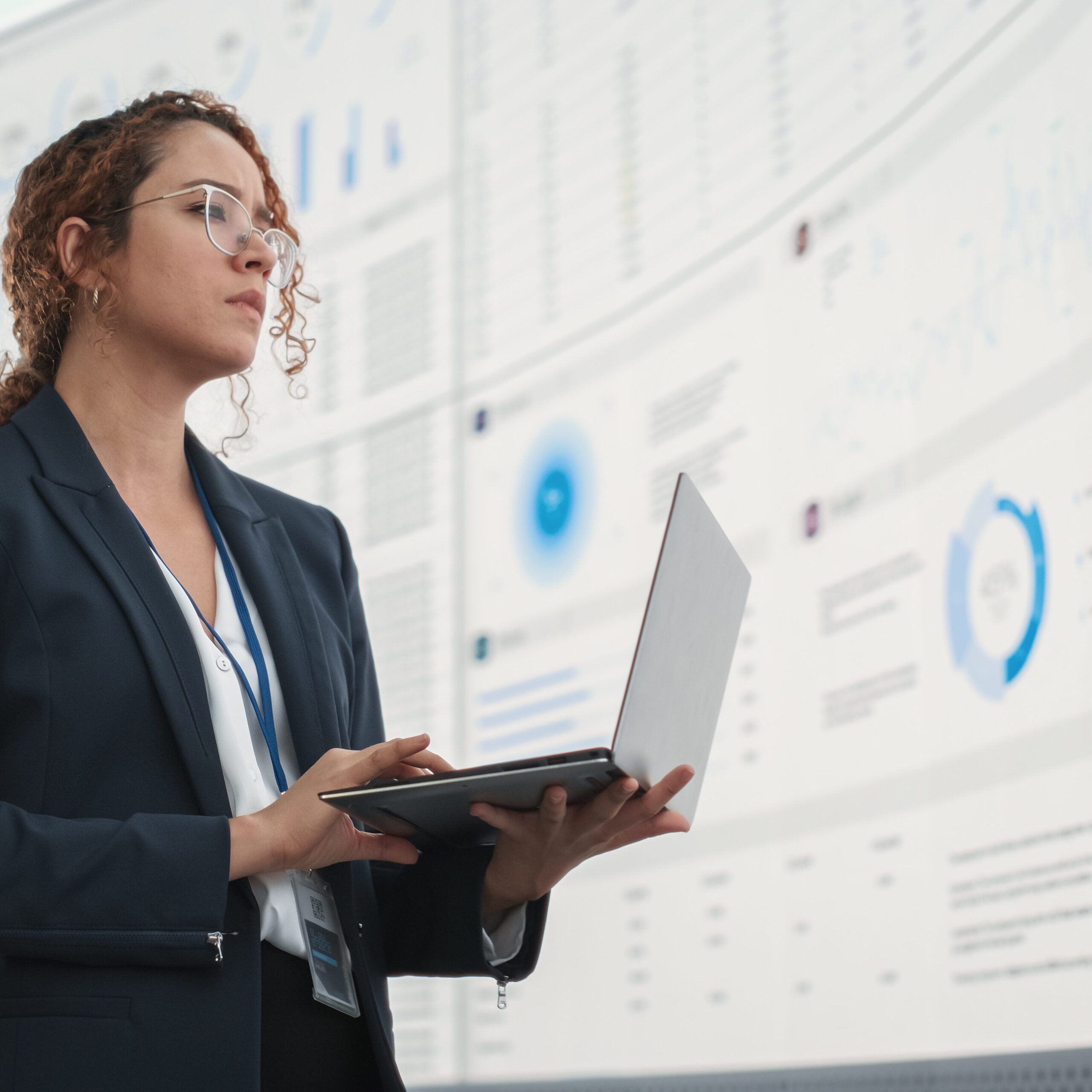 Successful Hispanic Businesswoman Holding Laptop Computer And Reviewing Financial Reports On Digital Screen In Monitoring Office. Female Leader Running Deep Learning, Artificial Intelligence Company.