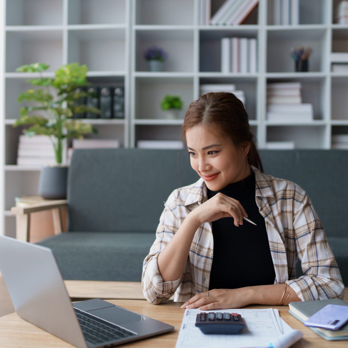 A woman sits at a table working on her laptop in a modern living room. She is smiling while holding a pen, with a calculator and documents in front of her.
