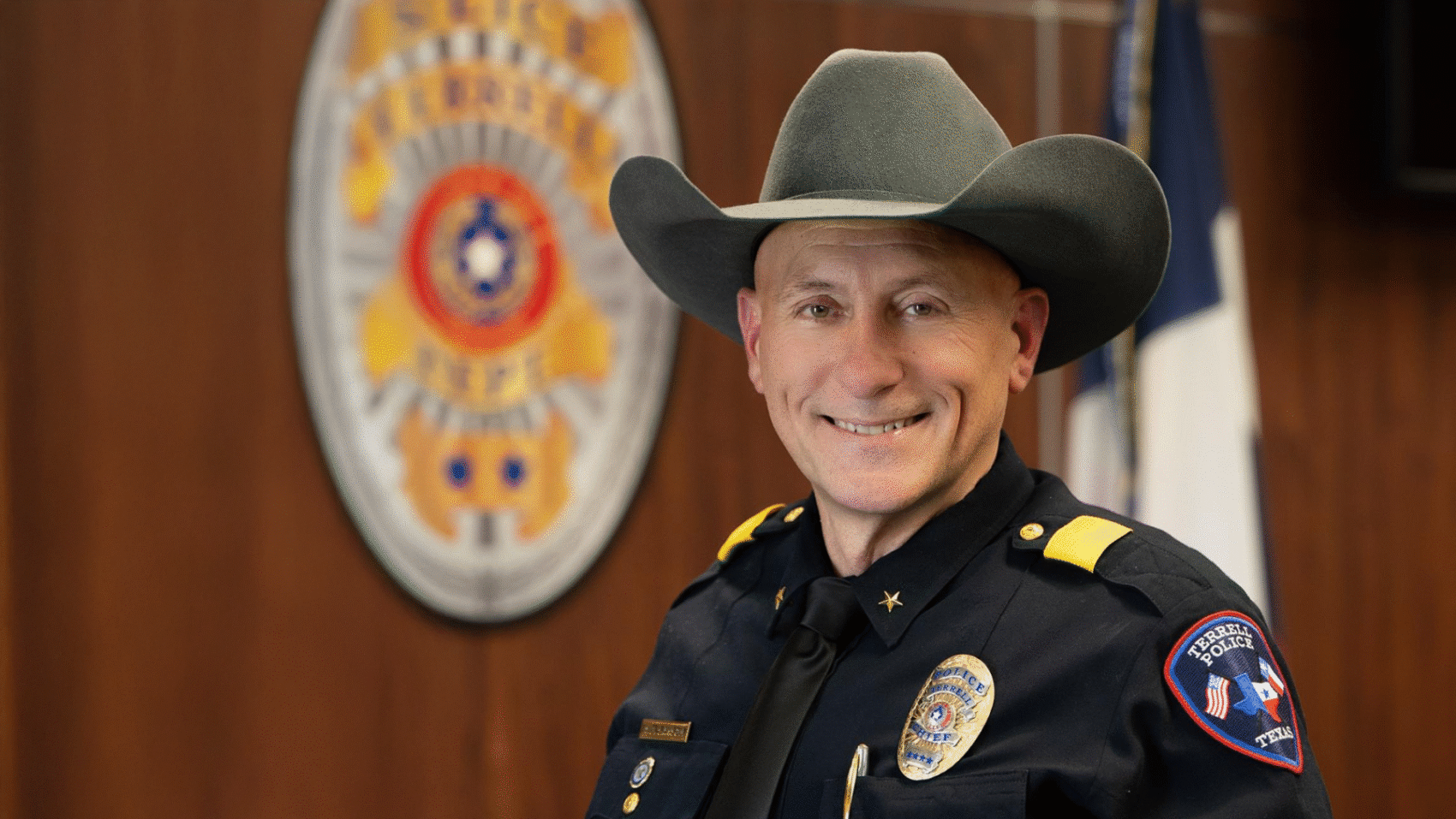 A man dressed in his police uniform and wearing his new chief's badge, against a woodgrain wall.