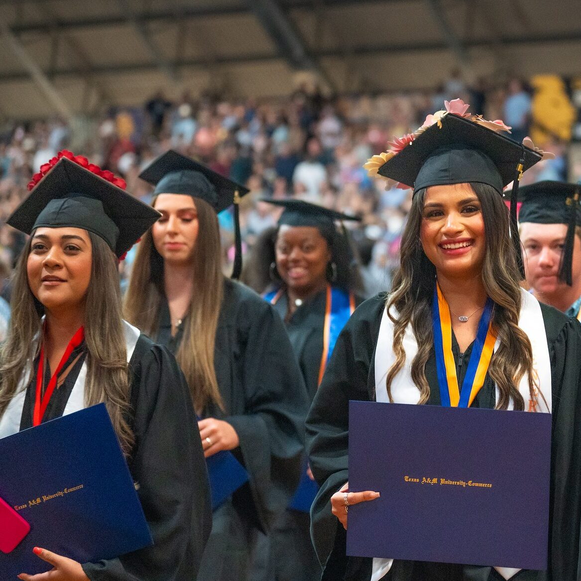 Graduates stand with their caps and gowns. Graduate in the foreground smiles with diploma in hand.