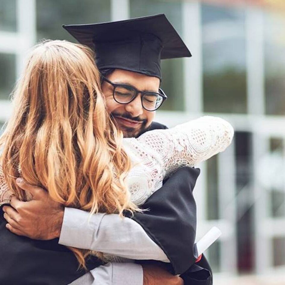 A graduate in a cap and gown happily embraces a woman with long blonde hair. They are outside, in front of a modern building with glass windows. The graduate holds a diploma in one hand, and appears joyful and proud. Sunlight shines gently in the background.