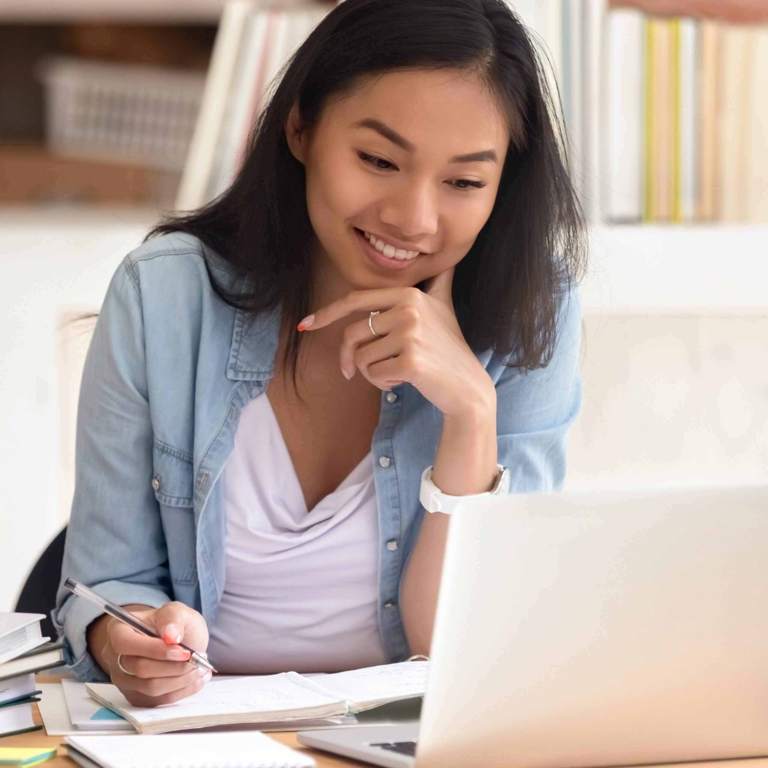Smiling asian undergraduate teen girl student study in library with laptop books doing online research for coursework, making notes for essay homework assignment, online education e-learning concept