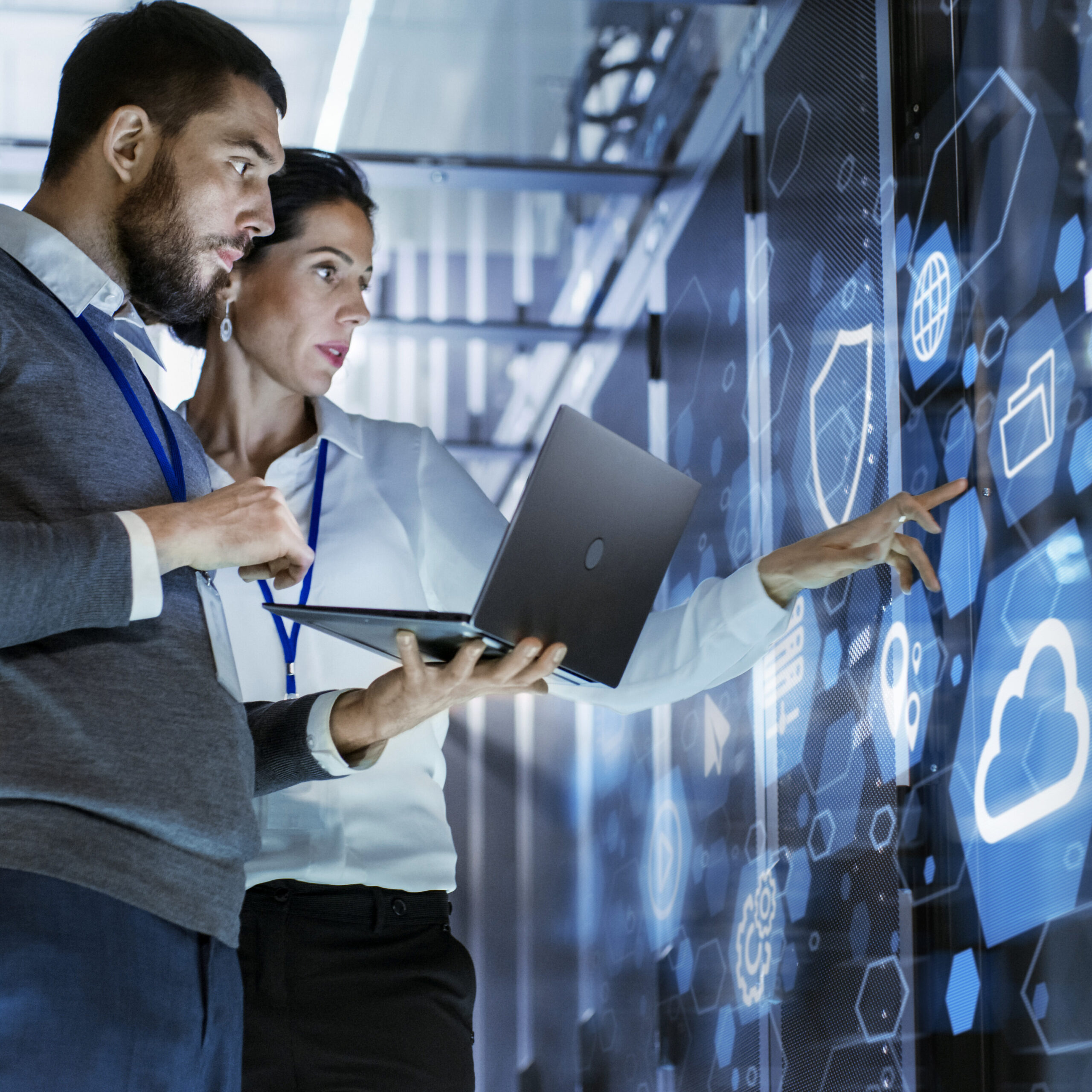 A female technology manager stands utilizing a touch screen console while her male counterpart holds a laptop at the ready.