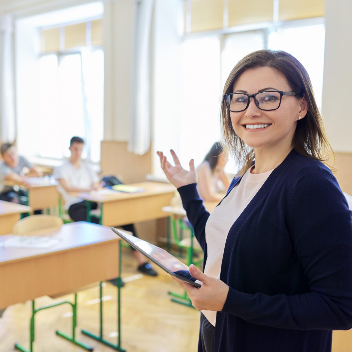 Portrait of female teacher at school at lesson, smiling middle-aged woman in glasses with digital tablet looking at camera, classroom with teenage students at desk background