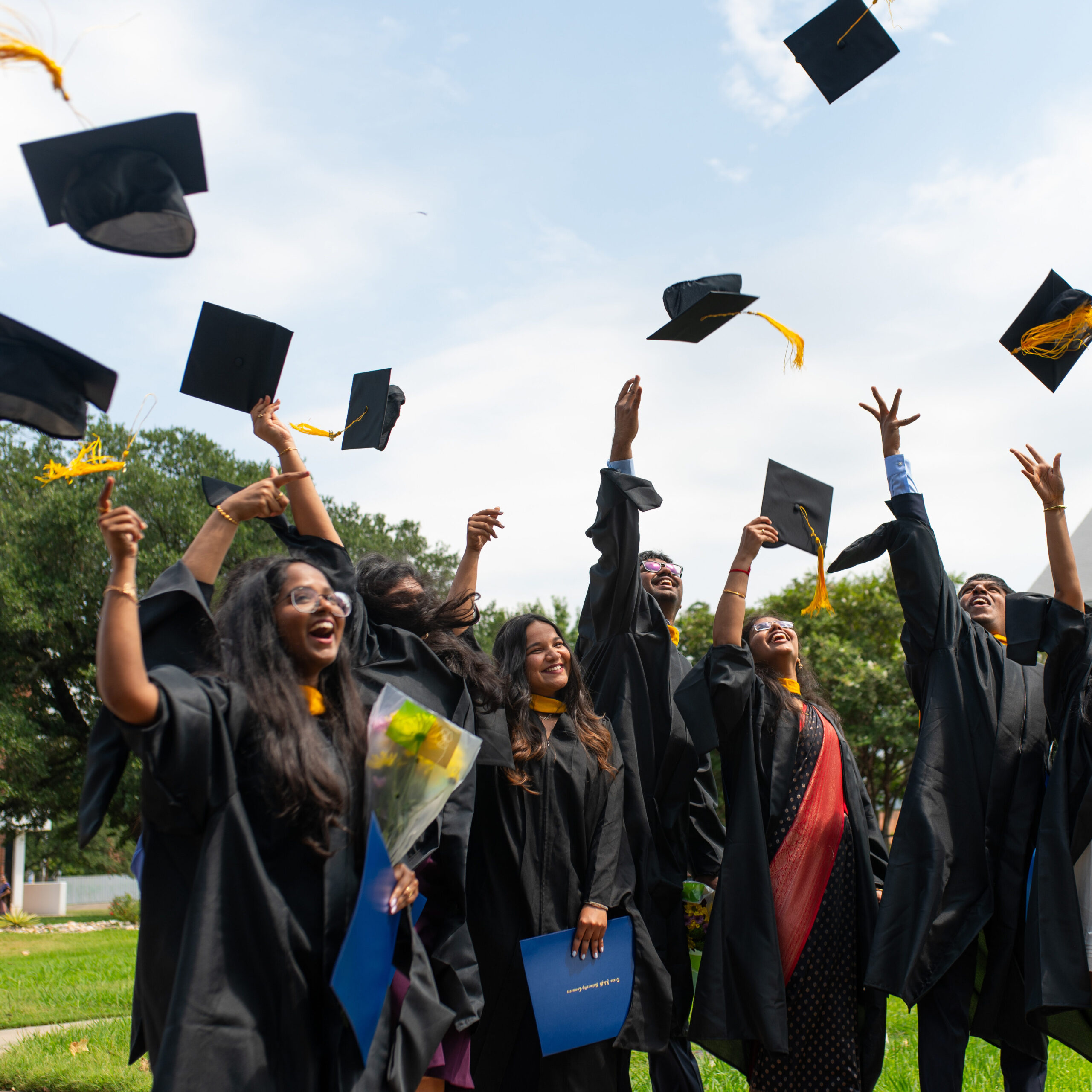Graduates in black gowns toss their caps into the air. Blue sky with clouds and green grass are in the background.