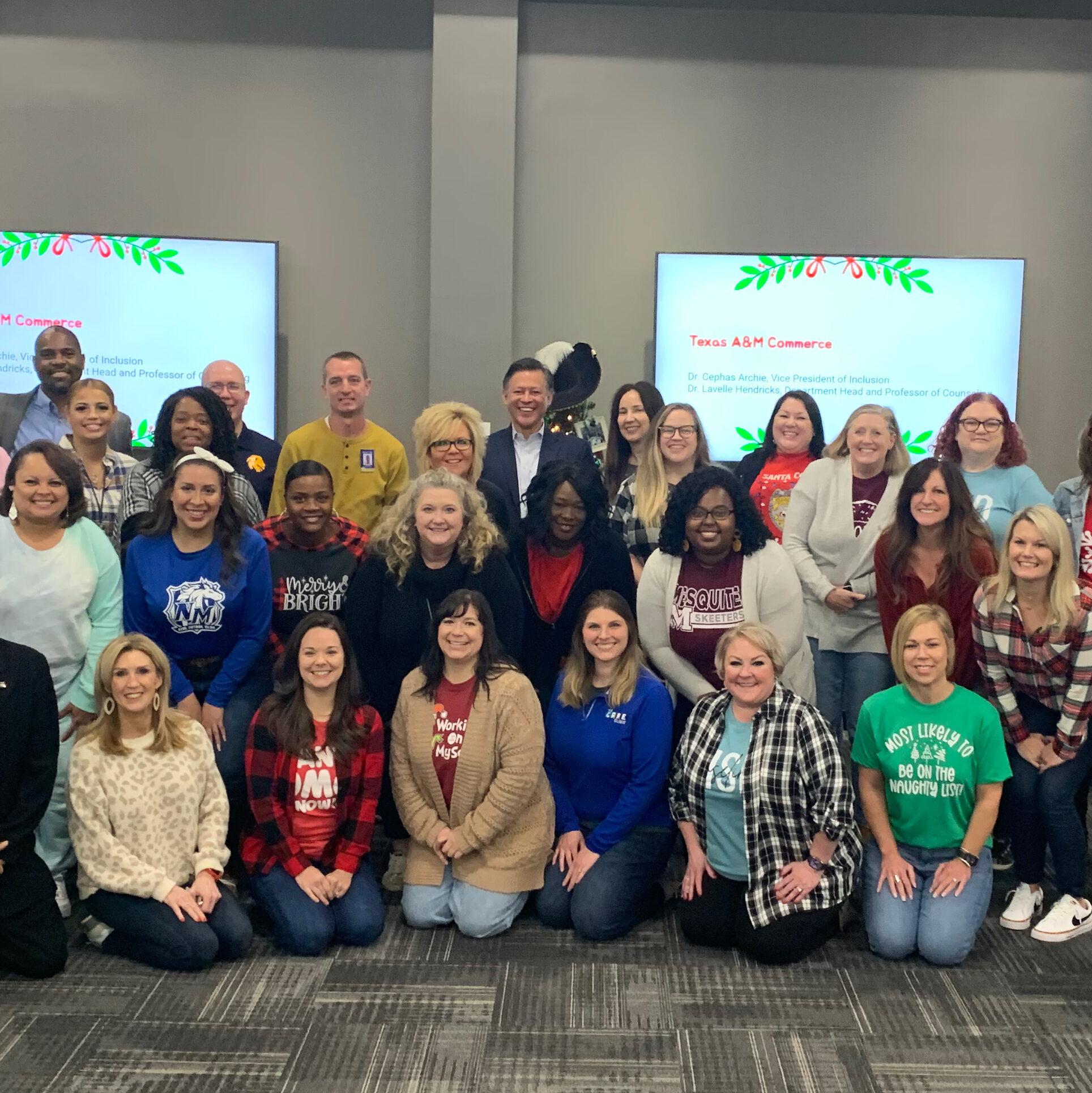 A group of adults standing and sitting in rows, while smiling for a group photo in a conference room. Two men in formal suits stand on the far left and right, while others wear casual attire, including festive holiday sweaters. Two screens display festive decorations behind them.