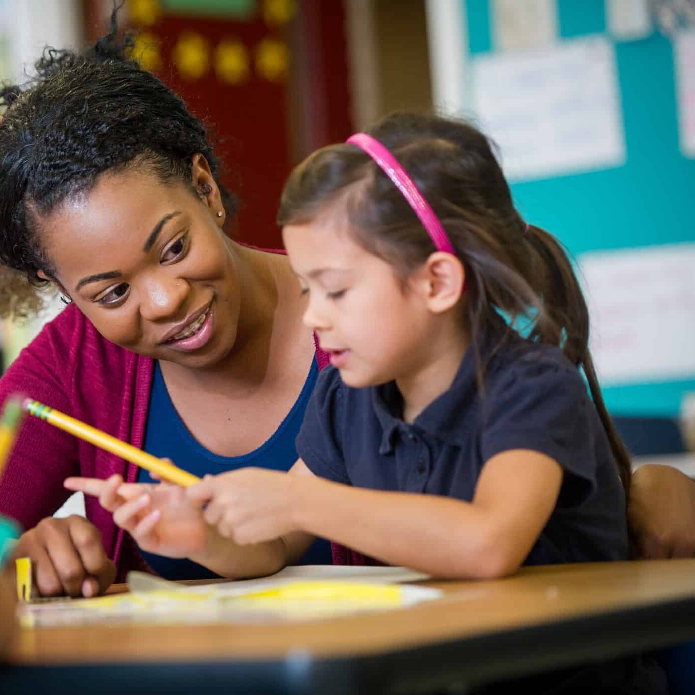 Female teacher with elementary age girl at school desk.