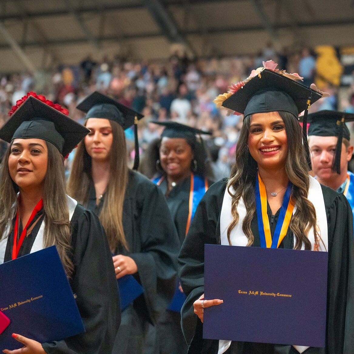 Graduates stand with their caps and gowns. Graduate in the foreground smiles with diploma in hand.