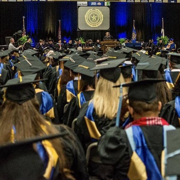 A large group of graduates in caps and gowns sit facing a stage during a commencement ceremony. The stage has officials and a podium, with a projection screen displaying a seal in the background. The venue is decorated with flags and banners.