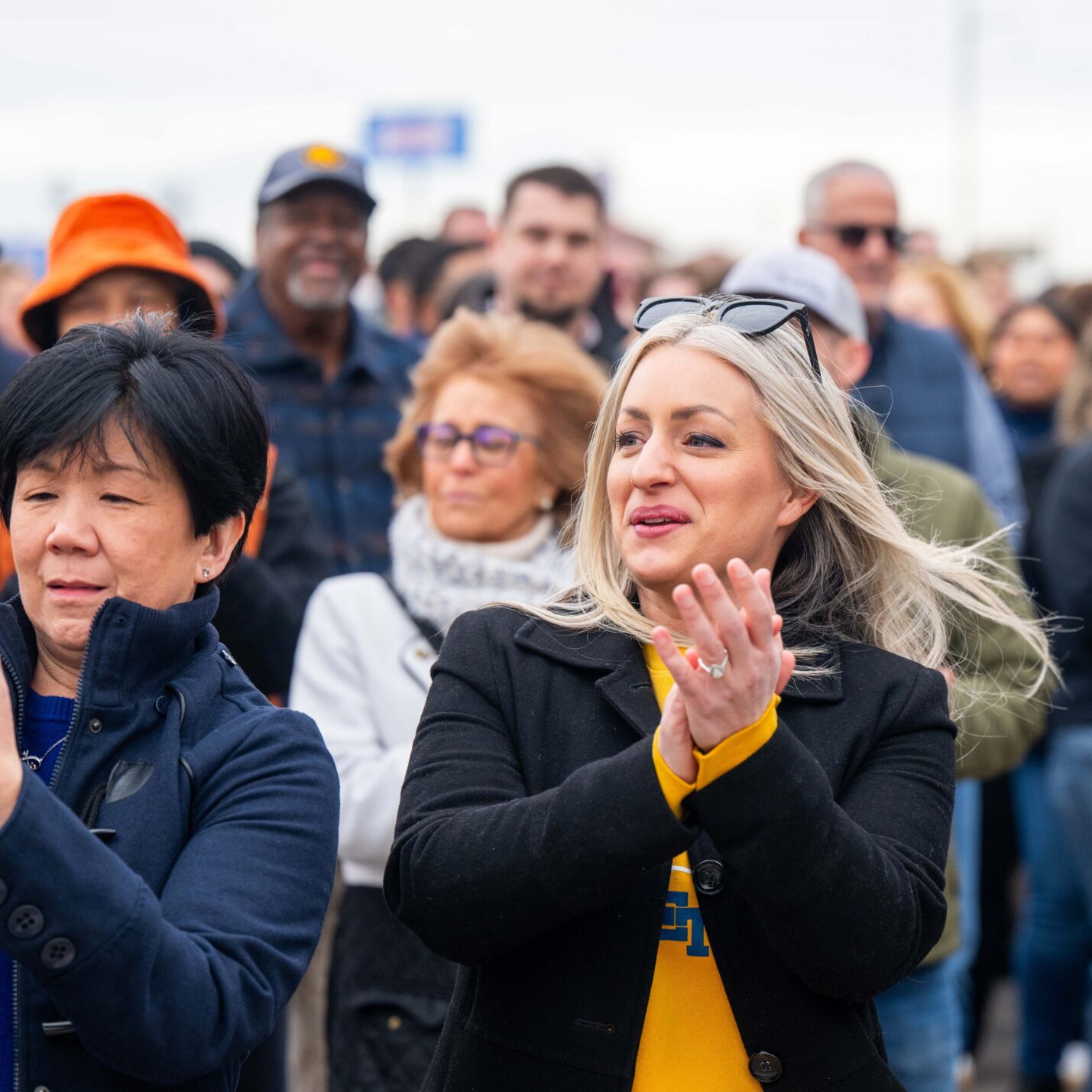 A crowd of people clap and smile. On the front row, two people take photos with their phones and one person smiles while clapping.