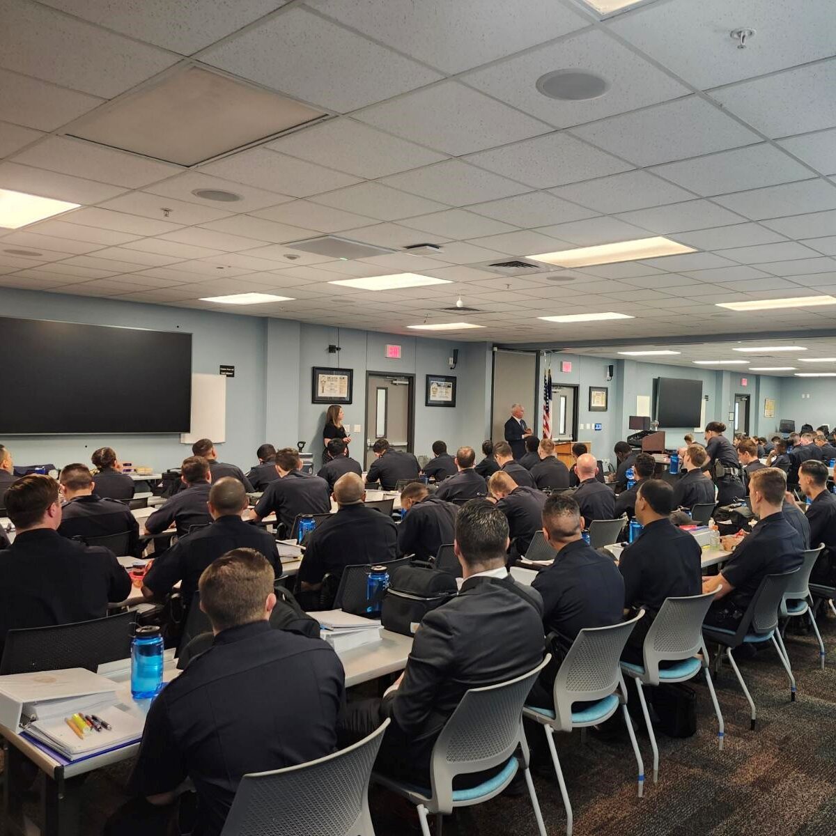 A multitude of police cadets sit at long tables listening to a presentation.
