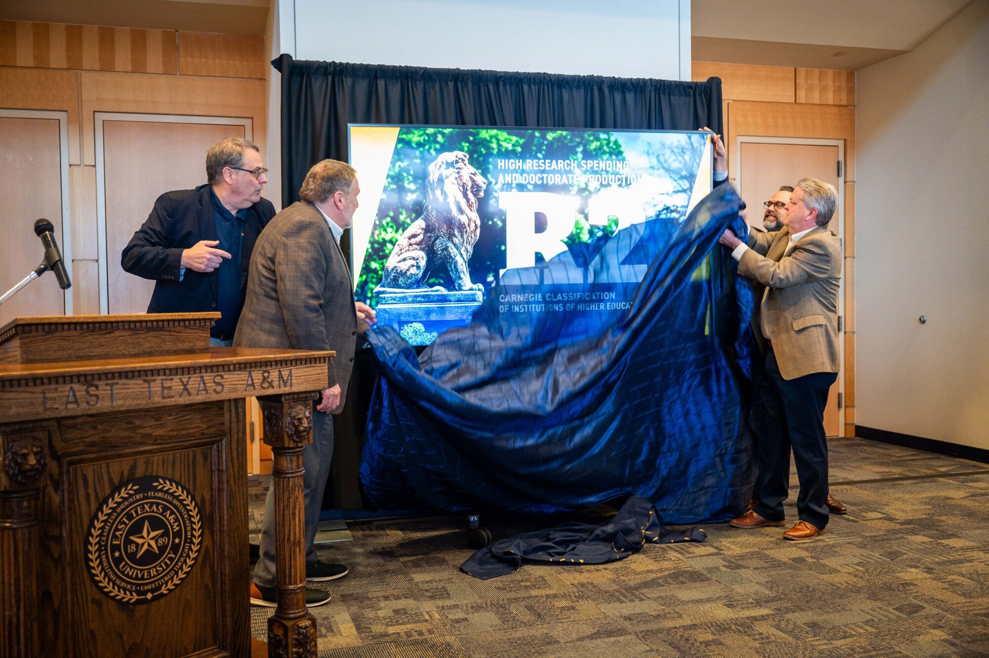 Two people on each side of a tv screen remove a large blue veil that is covering the screen. The screen says "R2" in large letters.